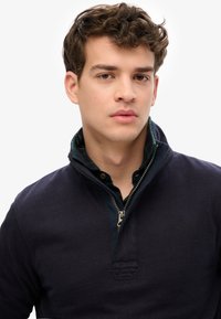 Marineblauwe pullover met een hoge kraag, ritssluiting en textuurstof; gedragen over een donker geruit shirt met knoopdetails.