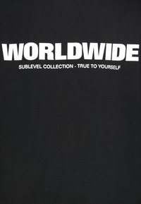 Zwarte katoenen t-shirt met grote witte tekst die "WORLDWIDE" leest, met daaronder "SUBLEVEL COLLECTION - TRUE TO YOURSELF".