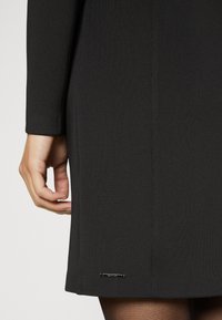Robe noire à la coupe ajustée, en tissu texturé, avec un accent de logo subtil près de l'ourlet. Gros plan de la manche et de la main.