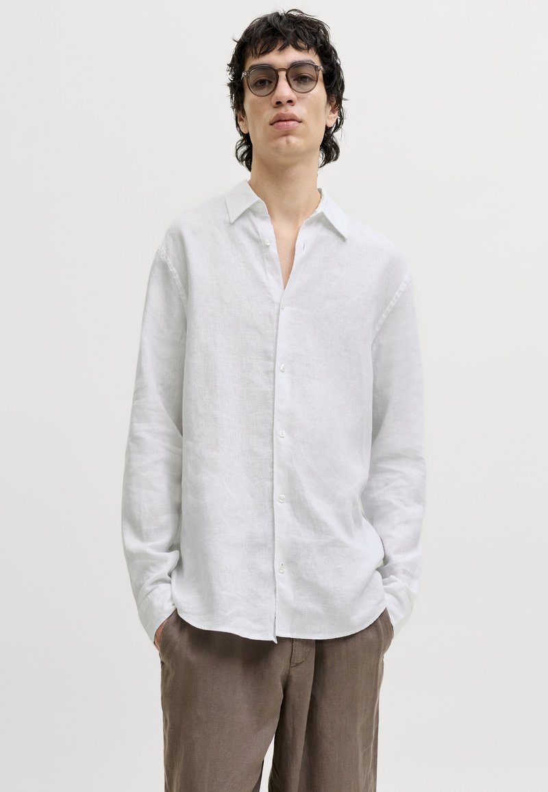 Personne aux cheveux courts et foncés et portant des lunettes, vêtue d'une chemise blanche à boutons et d'un pantalon marron, debout les mains dans les poches devant un fond uni.