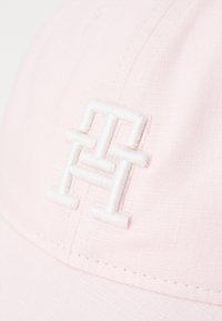 Rosa tygkeps med broderad vit logotyp. Har en böjd skärm och texturerat material. Närbilden framhäver sömnadsdetaljer.