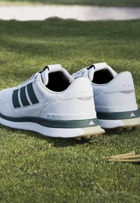 Λευκά και σκουροπράσινα παπούτσια γκολφ Adidas τοποθετημένα στο γρασίδι με ξύλινα tee γκολφ κοντά.