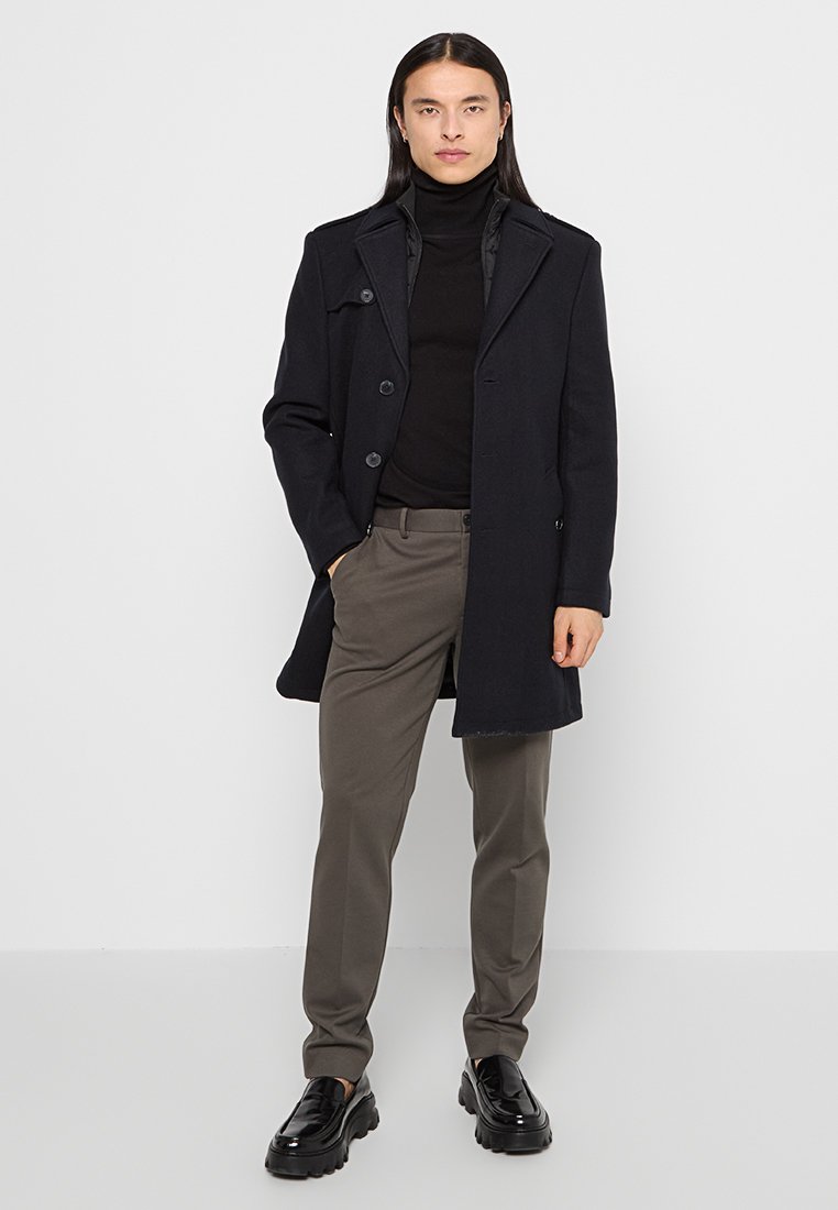Cappotto in misto lana blu navy con un design sartoriale, sei bottoni, revers a lancia, abbinato a un dolcevita nero e pantaloni grigi, completato con scarpe nere.
