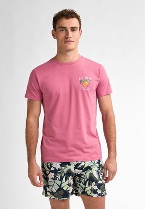 Roze katoenen T-shirt met "Key West" grafiek, gecombineerd met marineblauwe zwemshorts met bloemenprint met groene bladeren en witte bloemen.