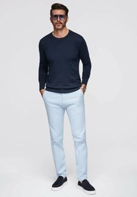 Marinefarbener Strickpullover, hellblaue Hose und dunkle Slippers. Das Modell steht lässig mit den Händen in den Taschen und trägt große Sonnenbrillen.
