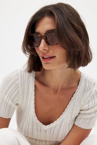 Skildpaddemønstrede solbriller med en tyk ramme. Modellen bærer en hvid tekstureret sweater med en dyb V-hals og korte puffede ærmer.