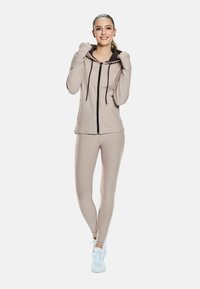 Young woman wearing a beige zip-up hoodie and matching leggings with white sneakers, posing with hands near the hood.