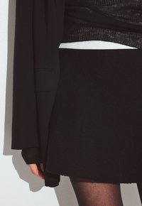 Suéter negro con tela texturizada superpuesto sobre una cintura blanca, acompañado de una falda negra acampanada y medias negras. Se ve una mano.