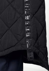 Černá prošívaná textilie s vertikálním lesklým černým pruhem, který nese opakující se text "KARL LAGERFELD".