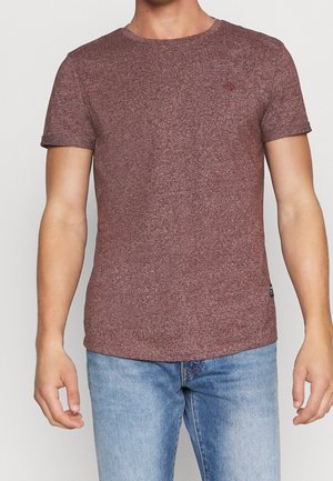 Man draagt een effen bordeauxrood T-shirt met korte mouwen en lichtblauwe jeans, staand met ontspannen armen tegen een witte achtergrond.