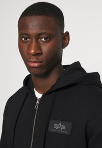 Black zip-up hoodie with a soft texture, featuring a front logo patch and drawstring hood. Strong focus on simple, functional design.