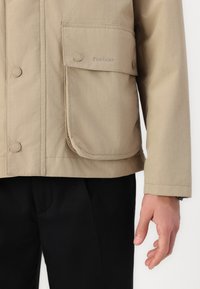 Beige dzseki gombos záródással, nagy elülső zsebbe és Barbour logóval. Simább anyag, szabott szabással, fekete nadrággal párosítva.