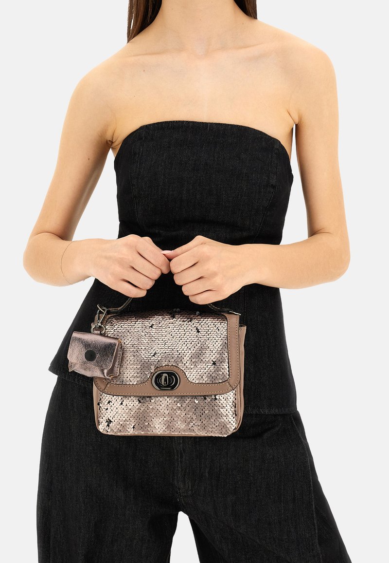 Donna in top senza spalline nero e pantaloni, che tiene in mano una piccola borsa beige con paillettes, dotata di un fermo frontale e una tasca attaccata.