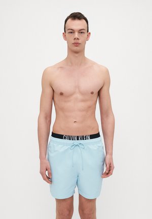 Svetlo modré plavecké šortky s pásom na stiahnutie a čiernym elastickým pásom s logom Calvin Klein. Hladká textúra, dizajn po kolená.
