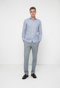 Hellblau kariertes Hemd, Slim Fit, mit langen Ärmeln. Kombiniert mit grauen Chinos und schwarzen Slippern, vor einem weißen Hintergrund stehend.