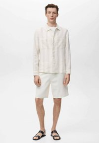 Witte, gestreepte linnen blouse met een kraag, twee voorzakken, gecombineerd met witte denim shorts en zwarte sandalen op een effen achtergrond.