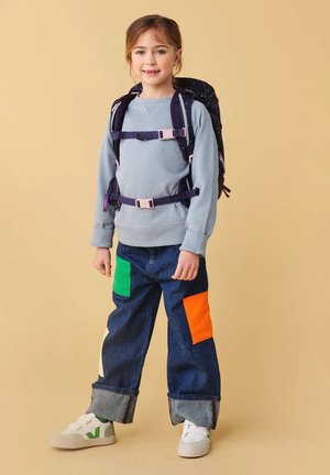 Navyfarbener Rucksack mit Sternen, grauer Sweatshirt, weite Jeanshose mit grünen und orangenen Patches und weiße Sneakers mit grünen Akzenten.