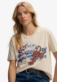 Beżowa bawełniana koszulka z kolorową grafiką homara i kwiecistymi akcentami; ma krótkie rękawy i okrągły dekolt, noszona z ciemnymi dżinsami.