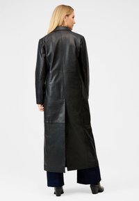 Lång svart läderrock med en slät yta, strukturerad krage och sidoflik. Bärs över vida byxor och klackstövlar. Minimala sömmar.