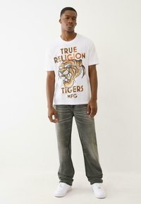 Młody mężczyzna w białej koszulce z grafika tygrysa i napisem "True Religion Tigers MFG", szarych dżinsach i białych trampkach, stojący na tle białym.