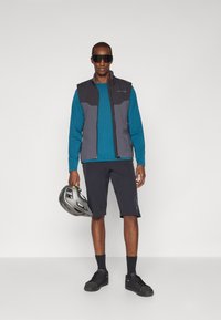 Sportliche Outfits mit einer grauen Weste mit schwarzen Akzenten, einem langen türkisfarbenen Shirt, schwarzen Shorts, schwarzen Schuhen und einem grauen Helm in der Hand.