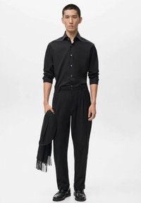 Camisa negra de manga larga, con frente abotonado, combinada con pantalones negros de talle alto y zapatos negros. Sostiene una bufanda negra.