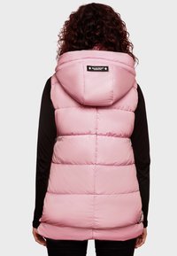Rosa pufferväst med huva, med ett quiltat mönster och en svart logotyp på baksidan, bärs över en långärmad svart topp.