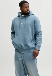 Sweat à capuche en coton bleu avec une poche avant et des cordons, arborant un logo blanc "JJ Studios" sur la poitrine. Porté avec un jean bleu clair.