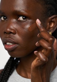 Un liquide brun appliqué sur la joue d'une personne à la peau foncée. La personne a des cheveux tressés et porte un haut gris clair. Texture de la peau naturelle visible.