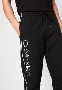Black sweatpants featuring white "Calvin Klein" text along the side, elastic waistband with drawstring, and contrasting side stripes.