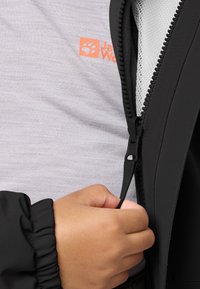Giacchetto nero con zip, indossato sopra una maglietta grigia chiara con un logo arancione. Una mano afferra il tirante della zip per chiuderla.