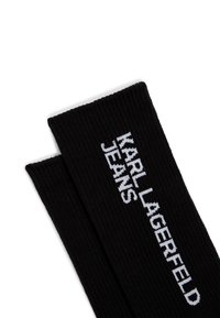Svarta ribbade strumpor med vit broderad text som läser "KARL LAGERFELD JEANS" längs sidan, med en slät och flexibel textur.
