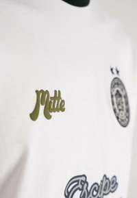 Țesătură albă cu text "Mitte" de culoare verde măsliniu, un emblema circulară întunecată cu două stele deasupra și text albastru cursiv parțial vizibil dedesubt.