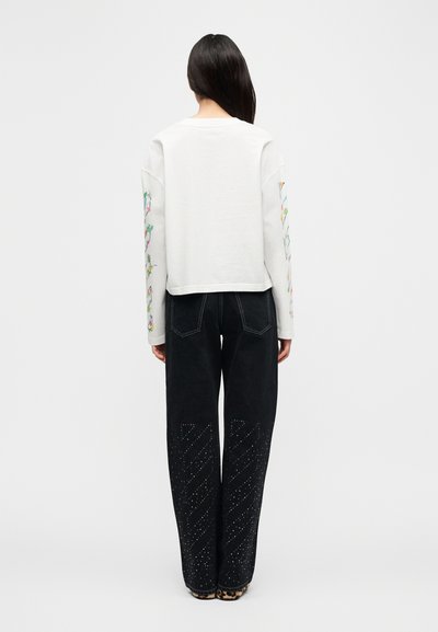 Λευκό πουκάμισο με μακριά μανίκια και κεντήματα με λουλούδια στα μανίκια, συνδυασμένο με μαύρο τζιν φαρδιάς γραμμής με διαγώνια διακοσμητικά καρφάκια στα πόδια.