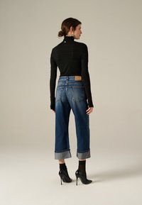 Donna di spalle che indossa un dolcevita nero a maniche lunghe, jeans blu con ampio risvolto, calze nere trasparenti, scarpe nere con tacco alto e orecchini d'argento.
