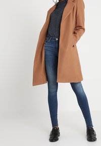 Brauner langer Mantel über einem dunkel gemusterten Oberteil, kombiniert mit blauen Skinny Jeans und schwarzen flachen Schuhen. Hat einen klassischen Kragen und eine taillierte Passform.