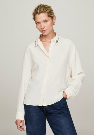 Camicia button-up crema con colletto a contrasto nero e maniche lunghe, abbinata a jeans in denim blu scuro. Tessuto morbido e vestibilità rilassata.