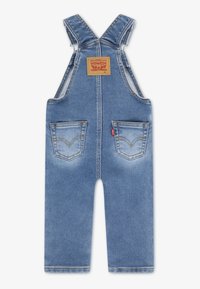Salopetă din denim bleu deschis cu bretele reglabile, o pată pătrată pe spate și două buzunare frontale decorate cu detalii cusute.