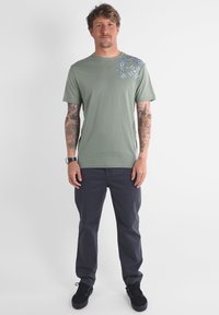 T-shirt en coton vert avec un motif floral bleu sur l'épaule gauche, assorti à un pantalon gris foncé et des baskets noires. Fond plat.