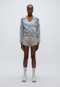 Ανοιχτό γκρι αδιάβροχο με σμικρυμένο μέση, συνδυασμένο με ανοιχτό μπεζ σορτς. Το μοντέλο φοράει λευκά αθλητικά παπούτσια και λευκές κάλτσες μέχρι το γόνατο.