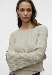 Lys beige, plisserte langermet topp med båtutringning og utsvingte mansjetter, laget av teksturert stoff. Modell med armene korslagt, nøytral bakgrunn.