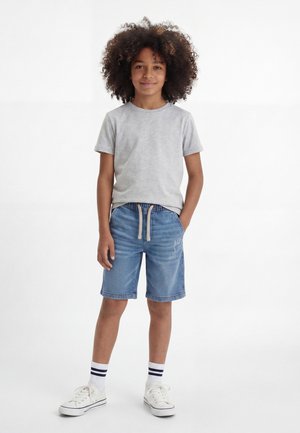 Dziecko z kręconymi włosami stoi, mając na sobie jasnoszarą koszulkę, niebieskie krótkie spodenki jeansowe ze sznurkiem, białe skarpetki w paski i białe trampki.