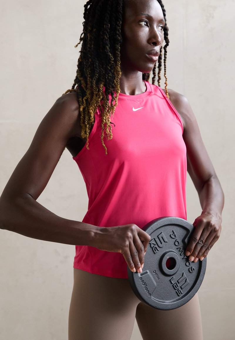Różowy sportowy top na ramiączkach z logo Nike, zestawiony z beżowymi legginsami. Osoba trzyma czarną płytę do ćwiczeń, prezentującą zarys mięśni.