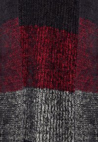 Tessuto lavorato a maglia con un motivo geometrico composto da blocchi di rosso scuro, nero e grigio, che mostra diverse texture e spessori.