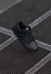 Juodas Nike sportbačius su tekstūruota odos viršumi, geltonais logotipo akcentais, perforuota pirštų dėžute ir plokščiais juodais raišteliais. Įrengtas guminiu padu.