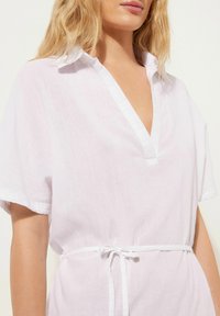 Vestido de lino blanco con escote en V, mangas cortas y cintura ajustada. Textura suave con un corte relajado y detalle de cuello en contraste.