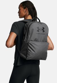 Szary plecak Under Armour o gładkiej fakturze, z wyraźnym logo i wieloma przegródkami. Prosty design z regulowanymi paskami.