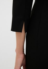 Schwarzer Kleidungsärmel mit einem Schlitz, strukturiertem Gewebe und einem klaren Design; hebt den Arm und die Hand vor einem neutralen Hintergrund hervor.