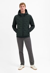 Donkergroene gewatteerde hoodie van een textuurstof, met ritssluiting en verstelbare koorden, gecombineerd met grijze broek en witte sneakers.