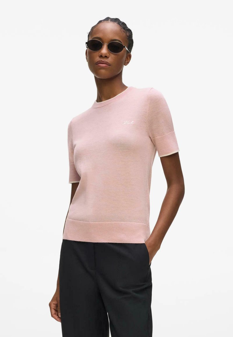 Femme portant un pull rose à manches courtes avec un logo, un pantalon noir et de petites lunettes de soleil ovales, debout avec une main dans la poche.